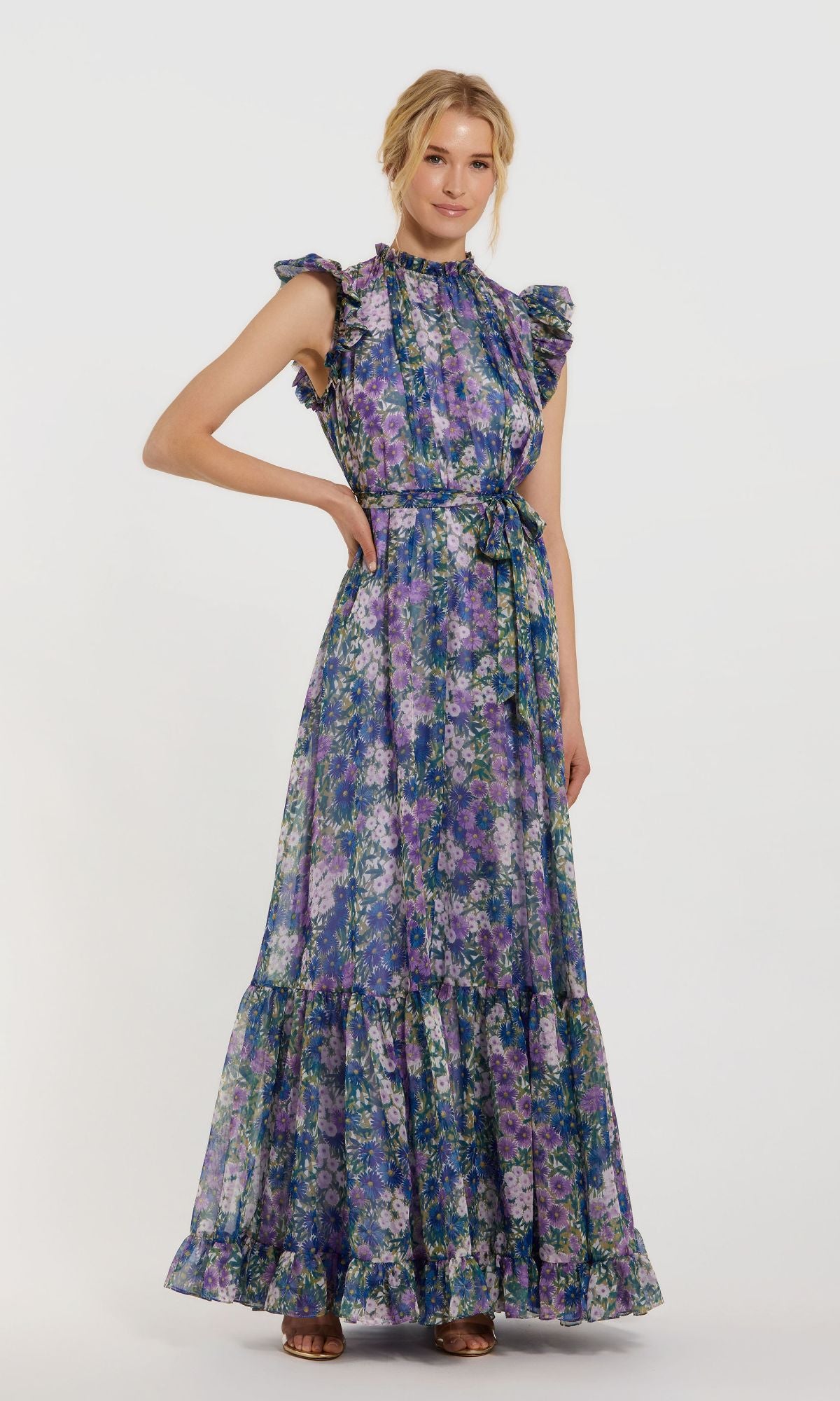 A woman wears the Mac Duggal 56116 Lilac Floral-Print Formal Gown, a floor-length, sleeveless dress with ruffled shoulders and a tie waist.